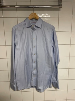 Ljusblå Eton - Säljer en stilren ljusblå Eton skjorta med klassisk krage och knappar framtill. Skjortan har lång ärm med manschett och är tillverkad i bomull för en bekväm känsla. Perfekt till både jeans och kostym.