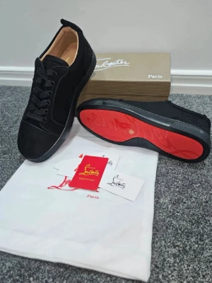 Svarta Louboutin sneakers med röd sula - Snygga svarta sneakers från Christian Louboutin med ikonisk röd sula. Skorna är låga och har ovandel i mocka med svarta skosnören och diskreta detaljer. Perfekta för dig som vill sticka ut med en lyxig och trendig look.