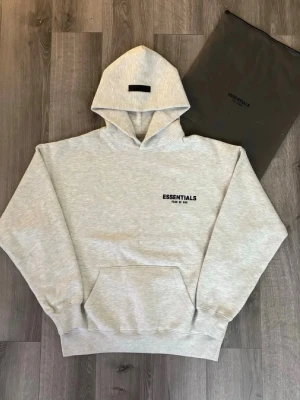 Grå Essentials Fear of God hoodie - Snygg ljusgrå hoodie från Essentials Fear of God med svart logga tryckt på bröstet. Klassisk oversized passform, stor huva och magficka framtill. Tillverkad i mjuk bomullsblandning som känns riktigt skön mot huden. Perfekt för en avslappnad streetwear-look. Anvönd tre gåbger