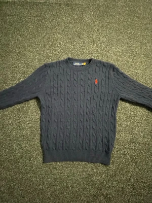 Mörkblå kabelstickad tröja Polo Ralph Lauren - Kabelstickad mörkblå tröja från Polo Ralph Lauren med klassisk röd logga broderad på bröstet. Tröjan har rund halsringning, långa ärmar och ribbade muddar vid ärmslut och nederkant. Perfekt för dig som gillar tidlös och stilren design. Lite liten i storlek 