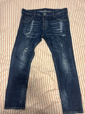Blå slitna jeans från Dsquared2 - Säljer ett par blå jeans från Dsquared2 med snygga slitningar och tvättade detaljer. Jeansen har klassisk femficksmodell, raka ben och en patch med logga bak i midjan. Perfekta för en avslappnad och trendig look. Klassiska dsq2 jeans