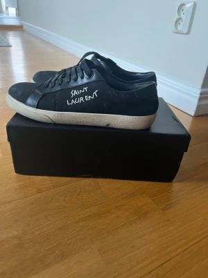 Svarta Saint Laurent sneakers - Svarta sneakers från Saint Laurent med vit text på sidan och klassisk låg profil. Skorna har svart läder och textil, vita sulor och svarta skosnören. Perfekta för en clean och stilren look.