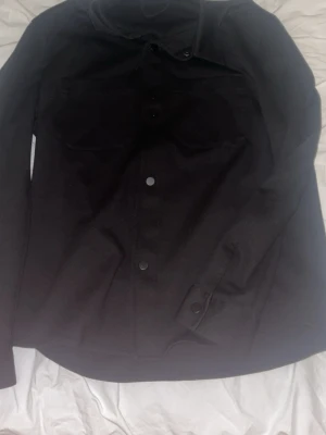 Svart jeansjacka i klassisk modell - Svart jeansjacka med knappar framtill och två bröstfickor. Jackan har en rak passform och är tillverkad i jeansmaterial. Perfekt för dig som vill ha en stilren och enkel look.