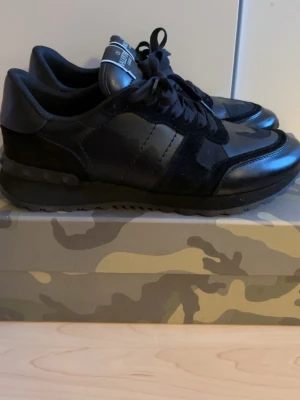 Valentino rockrunners - Säljer ett par exklusiva Valentino Rockrunner i svart. En ikonisk sneaker som kombinerar lyx och sportig design på ett stilrent sätt. Modellen är tillverkad i en mix av premiumläder, mocka och tekniskt tyg med Valentinos klassiska kamouflagedetaljer och den karaktäristiska nit-detaljen baktill. Skon är både bekväm och statement på samma gång – perfekt till allt från jeans och hoodie till en mer uppklädd look. Dom är använda fåtal gånger och är i topp skick. Låda och krease skydd tillkommer