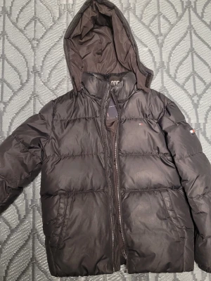 Svart pufferjacka från Tommy Hilfiger - Snygg svart pufferjacka från Tommy Hilfiger med huva och dragkedja framtill. Jackan har diskreta logodetaljer på bröstet och ärmen samt två sidofickor. Tillverkad i ett glansigt material som ger en modern look. Perfekt för kalla dagar.