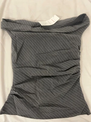 Grå randig offshoulder topp INDEFEIR - Snygg grå offshoulder topp från INDEFEIR med tunna vita ränder. Toppen har en tight passform och är perfekt för dig som gillar en stilren och trendig look. Materialet känns mjukt och stretchigt, vilket gör den bekväm att bära hela dagen.