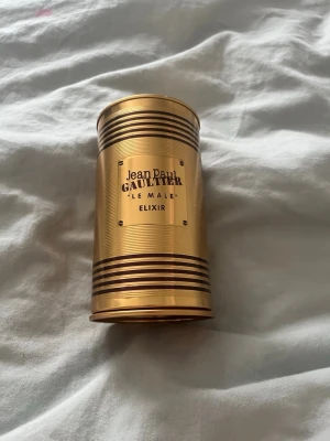 Jean Paul Gaultier Le Male Elixir - Snygg parfymförpackning i metall från Jean Paul Gaultier, modell Le Male Elixir. Guldfärgad cylinderformad burk med svarta ränder och präglad logga. Perfekt för dig som gillar exklusiv design och vill ha något unikt på parfymhyllan parfym fyller ej med den passar som en bra och snygg prydnad. 