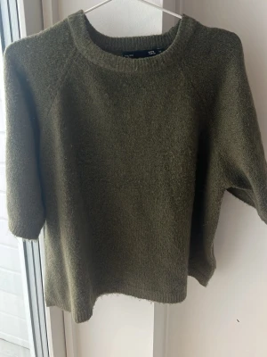 Olivgrön stickad tröja med halvlånga ärmar - Mysig olivgrön stickad tröja med rund halsringning och halvlånga ärmar. Tröjan har en enkel, rak passform och är tillverkad i ett mjukt material som passar perfekt för lager på lager. Stilren och lätt att matcha till olika outfits.