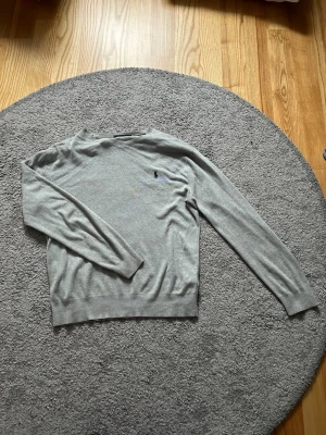 Grå stickad tröja från Ralph Lauren - Snygg grå stickad tröja från Ralph Lauren med klassisk marinblå logga broderad på bröstet. Tröjan har rund halsringning, långa ärmar och ribbade muddar. Perfekt till lager på lager och enkel att matcha med allt.