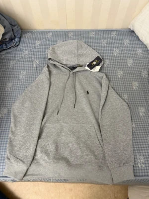 Grå hoodie från Polo Ralph Lauren - Snygg grå hoodie från Polo Ralph Lauren med klassisk huva och dragsnören. Tröjan har en liten broderad logga på bröstet och en stor magficka framtill. Tillverkad i mjukt bomullsmaterial som känns skönt mot huden. Perfekt för en avslappnad och stilren look.