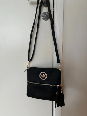 Svart axelväska från Michael Kors - Snygg svart axelväska från Michael Kors i konstläder med guldfärgade detaljer. Väskan har justerbar axelrem, dragkedja framtill och två dekorativa tofsar i läderimitation. MK-logga i metall på framsidan ger en lyxig touch.