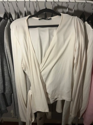 Vit omlottopp med knyt - Säljer en stilren vit omlottopp med långa ärmar och knyt i sidan. Toppen har en djup v-ringning och är gjord i mjuk bomullsmix som känns skön mot huden. Perfekt att matcha med jeans eller kjol för en clean look.