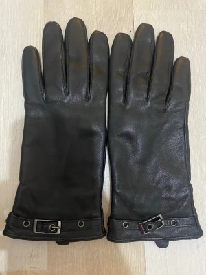 Svarta skinnhandskar med spänne - Läder handskar. Stilrena svarta skinnhandskar med dekorativt spänne och metall-detaljer vid handleden. Handskarna har klassisk form och är perfekta för kyliga dagar. Passar dig som vill ha en tidlös accessoar med lite edge.