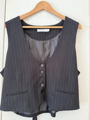 Kostymväst med smala ränder från Costes - Snygg mörkgrå/svart  kostymväst från Costes med tunna vita kritstrecksränder. Västen har fem knappar framtill, två dekorativa fickor och justerbart band i ryggen. Perfekt för en stilren och trendig look. Materialet är slätt och fodrat för extra komfort.
