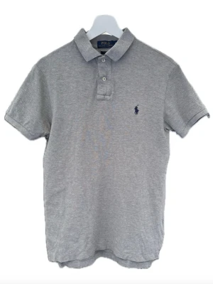 Grå pikétröja från Polo Ralph Lauren - Klassisk grå pikétröja från Polo Ralph Lauren med korta ärmar, krage och två knappar framtill. Tröjan har den ikoniska broderade loggan på bröstet och är tillverkad i mjuk bomull för en bekväm känsla. Storlek M men sitter som s