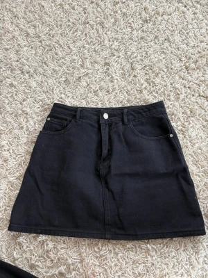 Svart jeanskjol med fickor - Snygg svart jeanskjol i klassisk A-linjeform med fem fickor och bälteshällor. Kjolen har knapp och dragkedja framtill och är gjord i ett stadigt denimtyg. Perfekt att matcha med sneakers eller boots för en cool look.