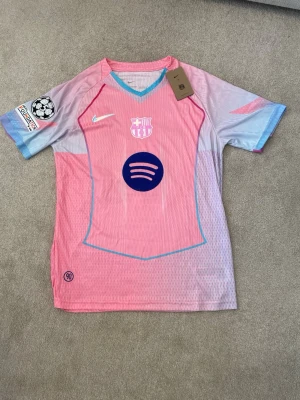 Barça rosa matchtröja Nike Yamal #10 - Säljer en Barcelona matchtröja i rosa och ljusblått från Nike med Champions League-märke på ärmen och Spotify-logga på bröstet. På ryggen står det Lamine Yamal och nummer 10. Tröjan är kortärmad och tillverkad i lätt polyester, perfekt för fotboll.