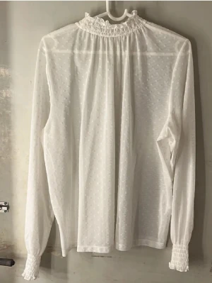 Vit transparent blus med volangkrage - Säljer en vit transparent blus med diskret prickmönster och volangkrage. Blusen har långa ärmar med smock vid ärmsluten och är gjord i ett tunt, lätt material som ger en luftig känsla. Perfekt för dig som gillar romantisk och trendig stil.