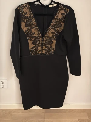 Svart spetsklänning från Oasis - Snygg svart kortklänning från Oasis med djup v-ringning och dekorativ spets i svart och beige framtill. Klänningen har långärmade ärmar och figurnära passform, perfekt för dig som vill sticka ut med elegans och stil.