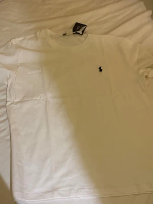 Vit t-shirt från Polo Ralph Lauren - Klassisk vit t-shirt från Polo Ralph Lauren med rund halsringning och den ikoniska lilla broderade loggan på bröstet. Tillverkad i mjuk bomull för en skön känsla och perfekt passform. Enkel och stilren design som funkar till allt.