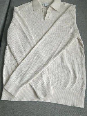 Vit stickad pikétröja från These Glory Days - Klassisk vit stickad pikétröja från These Glory Days med krage och v-ringad öppning. Tröjan har lång ärm, ribbade muddar och nederkant samt en diskret struktur i materialet. Perfekt för en clean och stilren look. Storlek S men sitter närmare XS.