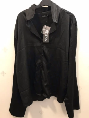 Svart satinpyjamas från Boohoo - Snygg svart pyjamas från Boohoo i mjukt satinmaterial. Setet består av en långärmad skjorta med krage och knappar samt matchande byxor med resår i midjan. Perfekt för chillkvällar hemma eller när du vill känna dig extra lyxig.
