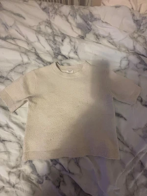 Beige stickad tröja från Kappahl - En enkel och stilren beige stickad tröja från Kappahl. Tröjan har korta ärmar, rund halsringning och ribbade kanter vid hals, ärmslut och nederkant. Perfekt för en avslappnad och trendig look.