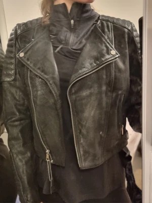 Svart biker skinnjacka med detaljer - Snygg svart skinnjacka i bikerstil med silvriga dragkedjor och tryckknappar. Jackan har vadderade partier på axlar och ärmar för en cool look. Kortare modell som passar perfekt till jeans eller klänning. Materialet är konstläder med matt finish.