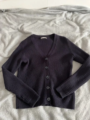 Mörklila ribbad kofta med knappar - Snygg mörklila ribbstickad kofta med v-ringning och fem stora knappar framtill. Långa ärmar och mjukt material som känns skönt mot huden. Perfekt att slänga över en t-shirt eller linne för en chill look.