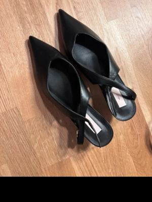 Svarta slingback med klack från Nelly - Stilrena svarta slingbacks med  från Nelly med spetsig v ringad och öppen häl. Skorna har en elegant slingback-rem och är tillverkade i ett slätt, konstläderliknande material. Perfekta för dig som gillar minimalistisk och trendig design. Oanvända och Nypris är 399 säljer säljer för 249