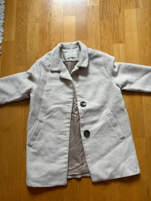 Ljusbeige rock från Zara Kids - Stilren ljusbeige rock från Zara Kids med två stora knappar framtill och snedställda fickor. Klassisk krage och helfodrad insida. Perfekt för dig som vill ha en enkel men snygg look under kallare dagar.