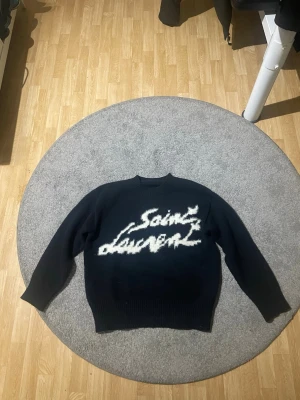 Svart stickad Saint Laurent tröja - Säljer en svart stickad tröja från Saint Laurent med vit, fluffig logotyp framtill. Tröjan har rund halsringning, långa ärmar och ribbade muddar vid ärmslut och nederkant. Perfekt för dig som gillar streetstyle och exklusiva märken.