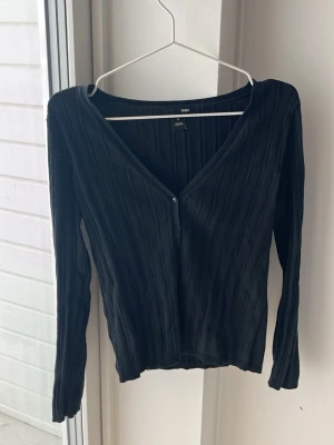 Svart ribbad cardigan från H&M - Säljer en svart ribbad cardigan från H&M med djup v-ringning och knäppning framtill. Tröjan har lång ärm och är tillverkad i ett mjukt, stretchigt material som sitter snyggt på kroppen. Perfekt att styla med jeans eller kjol.