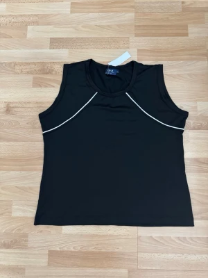 Ny. Svart ärmlös linnetopp med vit piping - Snygg svart linnetopp med vit piping längs bröstet. Toppen är ärmlös och har en rund halsringning. Perfekt för en clean och sportig look. Materialet känns mjukt och stretchigt. Bröst 110cm