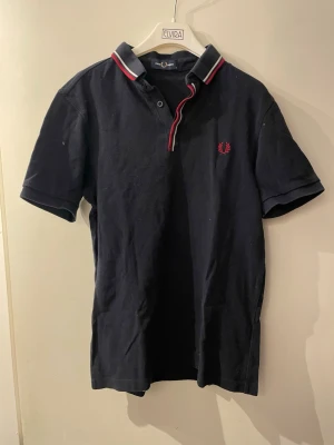 Marinblå piké från Fred Perry - Snygg marinblå pikétröja från Fred Perry med korta ärmar och klassisk krage. Kragen har röd och vit randig detalj och loggan är broderad i rött på bröstet. Perfekt för en stilren och sportig look.