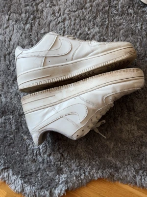 Vita Nike Air Force 1 sneakers - Klassiska vita Nike Air Force 1 sneakers i lågt utförande. Skorna har en stilren design med perforerad tå, vita skosnören och den ikoniska swoosh-loggan på sidan. Tillverkade i läder med gummisula för bra komfort och streetkänsla.