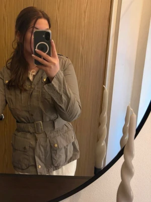 Barbour jacka - Barbour International jacka med bälte i midjan och flera fickor framtill. Jackan har en klassisk design med knappar och en hög krage. Ärmarna har justerbara manschetter med knappar. En logotyp finns på vänster ärm. Rökfritt hem