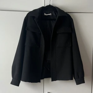 Svart kappa med fickor - Stilren svart kappa från ellos i ullmaterial med två stora bröstfickor och klassisk krage. Kappan har raka ärmar med knappar vid ärmslut och en enkel, boxig passform. Perfekt för att skapa en cool och tidlös look. OBS!! Den är avklippt längst ner och innertyget i ryggen är trasigt, men inget som syns när man har på sig den. 
