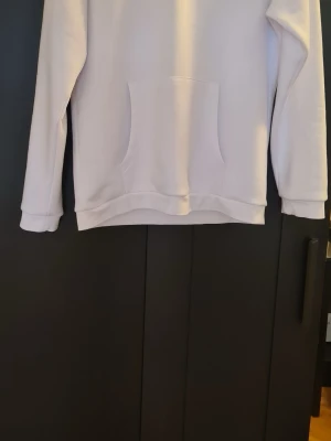 Vit hoodie från sail racing - En stilren vit hoodie med klassisk magficka och ribbade muddar vid ärmslut och nederkant. Hoodien har en avslappnad passform och är tillverkad i mjukt material, perfekt för en chill och enkel look.