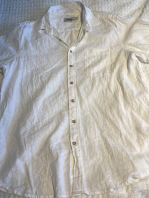 Vit kortärmad skjorta Dressmann - Klassisk vit skjorta från Dressmann i bomull med korta ärmar och bröstficka. Skjortan har knappar framtill och en avslappnad passform, perfekt till sommaren.