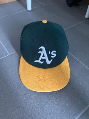 Oakland Athletics keps New Era - Snygg keps från New Era med Oakland Athletics A's-logga broderad framtill. Kepsen är mörkgrön med gul skärm och gul knapp på toppen. Klassisk basebollmodell med rak skärm och broderade ventilationshål. Perfekt för dig som gillar sportig stil.