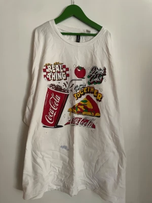 Vit Coca-Cola t-shirt med pizza-tryck - Säljer en vit t-shirt från Coca-Cola med färgglatt tryck av en colaburk, pizza, tomat och texten 'Real Thing' och 'Drink Coke'. T-shirten har rund hals och korta ärmar. Perfekt för dig som gillar retro och streetstyle vibes. (En fläck under trycket)