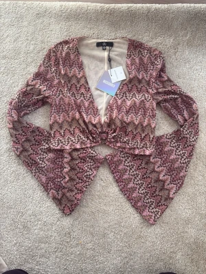 Chevronmönstrad topp med utsvängda ärmar - Säljer en rosa och brun chevronmönstrad topp från Missguided. Modellen har djup ringning, O-ring framtill och långa utsvängda ärmar. Materialet är mjukt och stretchigt, perfekt för en trendig look.