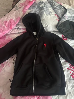 Svart hoodie från Ami Paris med dragkedja - Svart hoodie från Ami Paris med dragkedja framtill och klassisk huva. På bröstet sitter Ami:s ikoniska röda hjärta med A. Hoodien har känguruficka och ribbade muddar. Perfekt för en chill och stilren look.