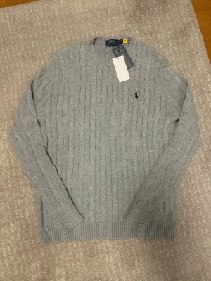 Ljusgrå kabelstickad tröja Polo Ralph Lauren - Snygg ljusgrå kabelstickad tröja från Polo Ralph Lauren med rund halsringning och diskret broderad logga på bröstet. Tröjan har ribbade muddar vid ärmslut och nederkant, perfekt för en stilren och klassisk look.
