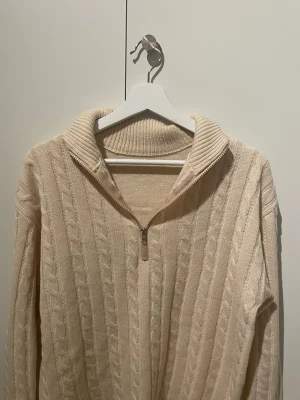 Beige kabelstickad tröja med halvzip - Kabelstickad beige tröja med halvzip och hög krage. Tröjan har långa ärmar och ribbade muddar vid ärmslut och nederkant. Perfekt för lager-på-lager och en chill look.