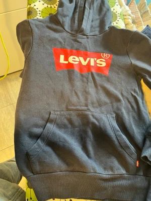 Svart Levi's hoodie med logga - Cool svart hoodie från Levi's med stor röd och vit logga på bröstet. Klassisk passform med huva och känguruficka framtill. Ribbstickade muddar vid ärmslut och nederkant. Perfekt för dig som gillar streetwear och ikoniska märken.