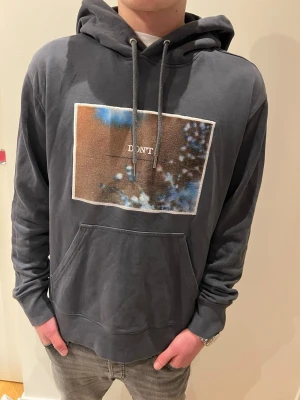 Mörkgrå hoodie J.Lindeberg  - Snygg mörkgrå hoodie med stor huva och dragsnören. Framtill finns ett rektangulärt print i blått, brunt och vitt med texten 'DON'T' i mitten. Hoodien har en klassisk känguruficka och är tillverkad i mjukt bomullsmaterial.