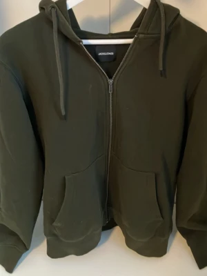 Mörkgrön hoodie från Jack & Jones - Snygg mörkgrön hoodie från Jack & Jones med dragkedja framtill och två stora fickor. Tröjan har huva med dragsko och ribbade muddar vid ärmslut och nederkant. Perfekt för en avslappnad och stilren look.