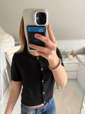 Kortärmad tröja - Snygg svart kortärmad kofta med pärlknappar framtill. Modellen är croppad och har rund halsringning. Perfekt att styla med jeans eller kjol för en trendig look. Materialet ser ut att vara mjukt och lättstickat.